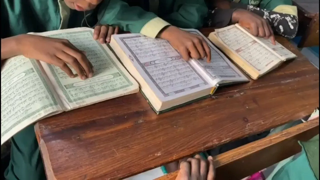 Élèves en uniforme lisant le Coran, livres ouverts sur une table en bois (seules les mains sont visibles)